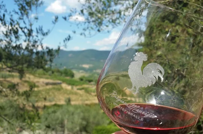 A glass of red wine etched with a black rooster held up against vineyard hills